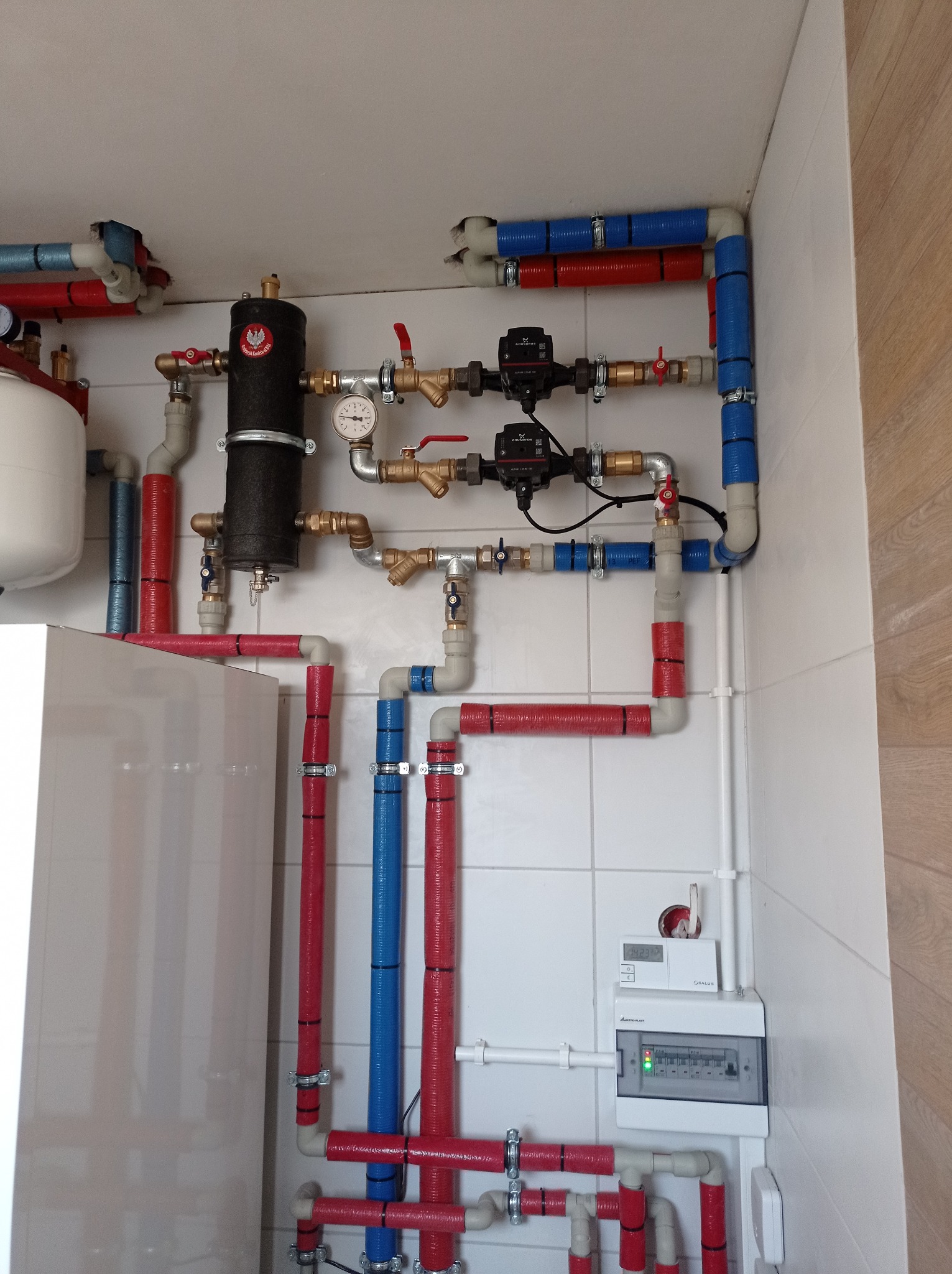 Instalacja grzewcza z rurami w kolorze czerwonym i niebieskim, zaworami, pompami obiegowymi i zbiornikiem wyrównawczym w kotłowni w Skierniewicach, widoczny również panel sterowania.