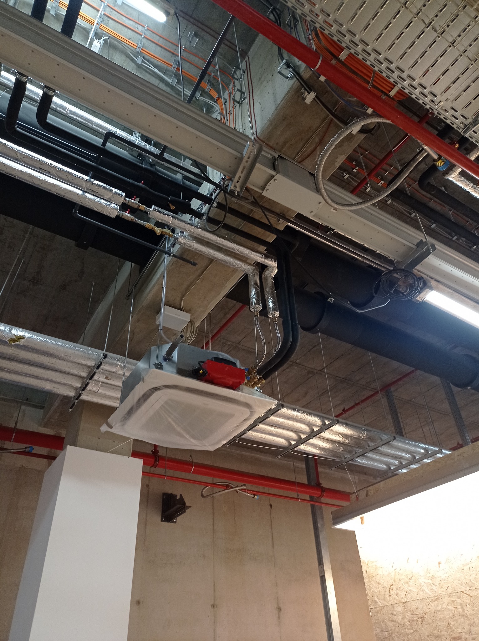 Montaż sufitowej jednostki klimatyzacyjnej w nowym budynku, widoczne rury i instalacje pod sufitem, jednostka zabezpieczona folią.