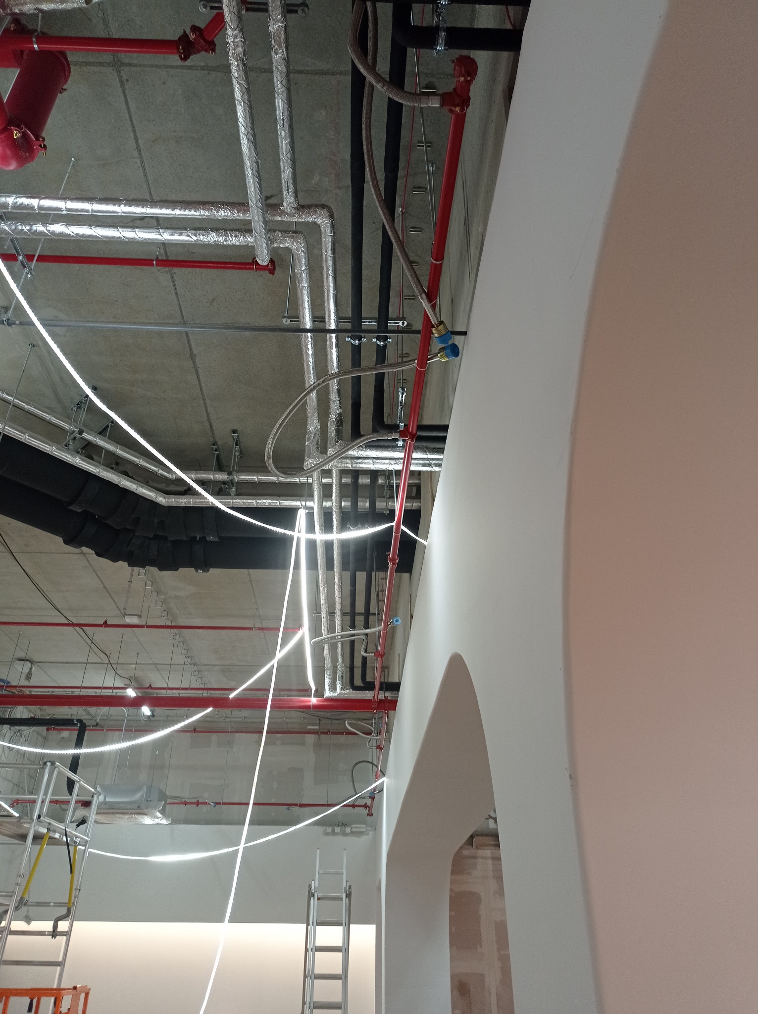 Widok na sufit w trakcie montażu instalacji: rury w izolacji aluminiowej, czerwone rury instalacji przeciwpożarowej, czarne rury wentylacyjne, białe przewody elektryczne, drabiny monterskie, łukowe...