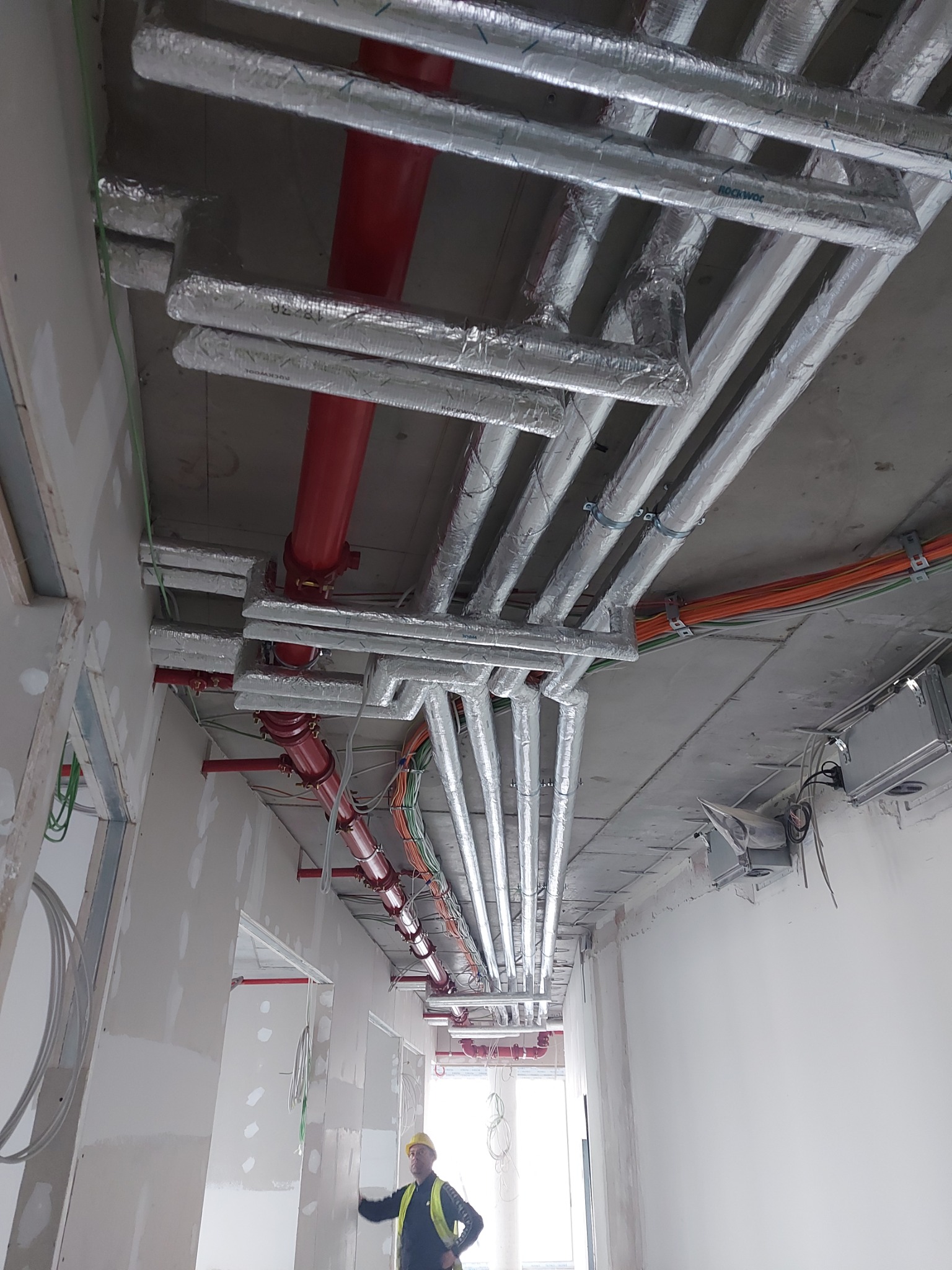 Widok instalacji wentylacyjnej i przeciwpożarowej pod sufitem w budynku w trakcie wykończenia, widoczne rury izolowane folią aluminiową, czerwone rury przeciwpożarowe, kable i pracownik w kasku.
