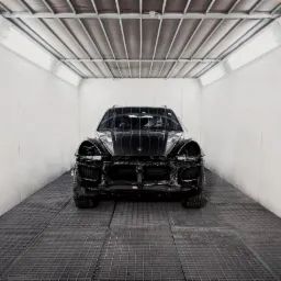 Czarny samochód osobowy, prawdopodobnie SUV, bez przedniego zderzaka i reflektorów, w jasnym pomieszczeniu z kratowaną podłogą, prawdopodobnie przygotowywany do naprawy karoserii.