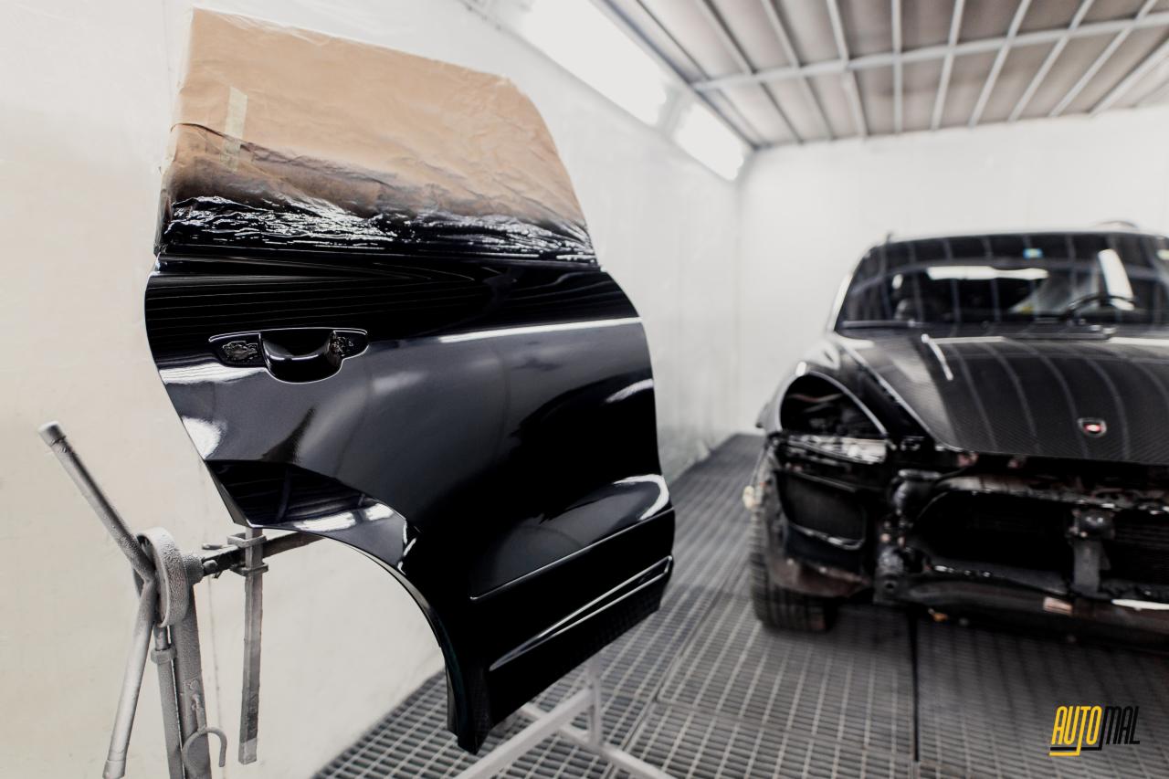 Czarny błyszczący element karoserii, prawdopodobnie drzwi, w trakcie lakierowania, zabezpieczony papierem, na stojaku w lakierni, z widocznym fragmentem samochodu w tle.