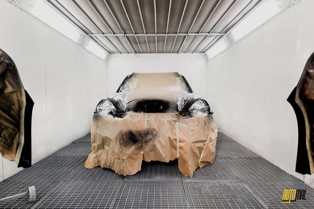 Samochód zabezpieczony folią i papierem w kabinie lakierniczej, przygotowany do malowania, widoczne czarne elementy karoserii i metalowa kratka podłogi.