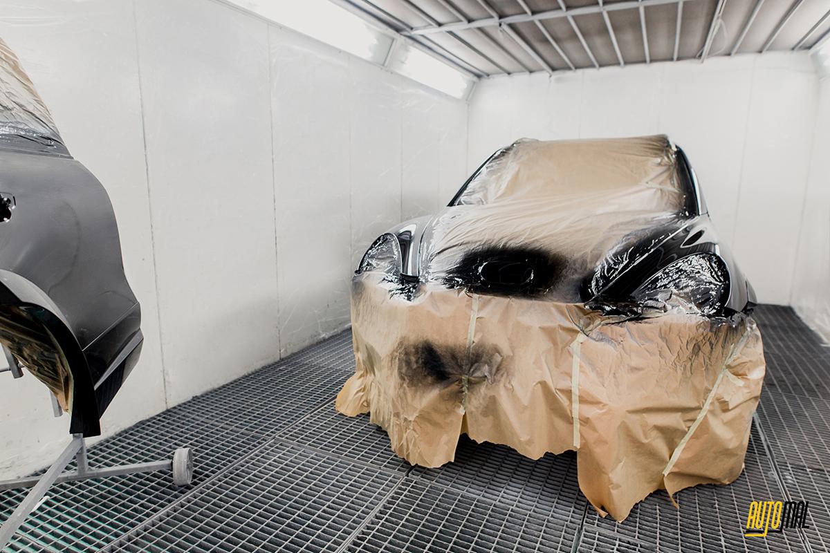 Przygotowane do lakierowania auto w kabinie lakierniczej, z zakrytymi elementami i świeżo pomalowanym błotnikiem na stojaku obok.