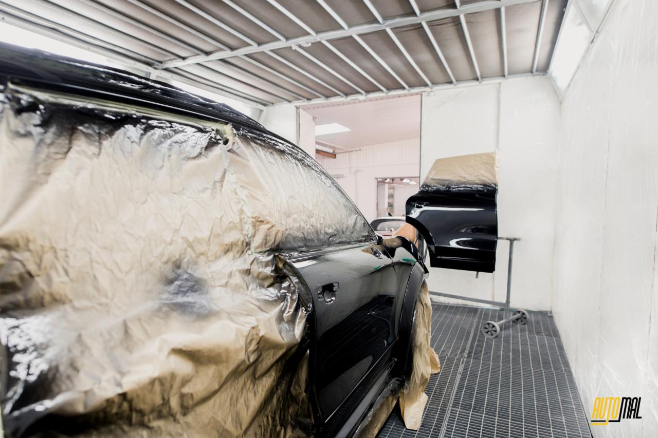 Przygotowanie czarnego samochodu do lakierowania w kabinie lakierniczej, z użyciem folii ochronnej. Widoczny fragment drugiego pojazdu w tle i logo firmy w prawym dolnym rogu.