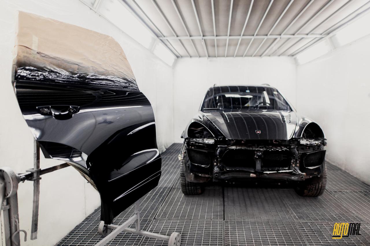 Czarny samochód w trakcie naprawy blacharsko-lakierniczej w kabinie lakierniczej, zdemontowane elementy karoserii, widoczny element karoserii na stojaku przygotowany do lakierowania.