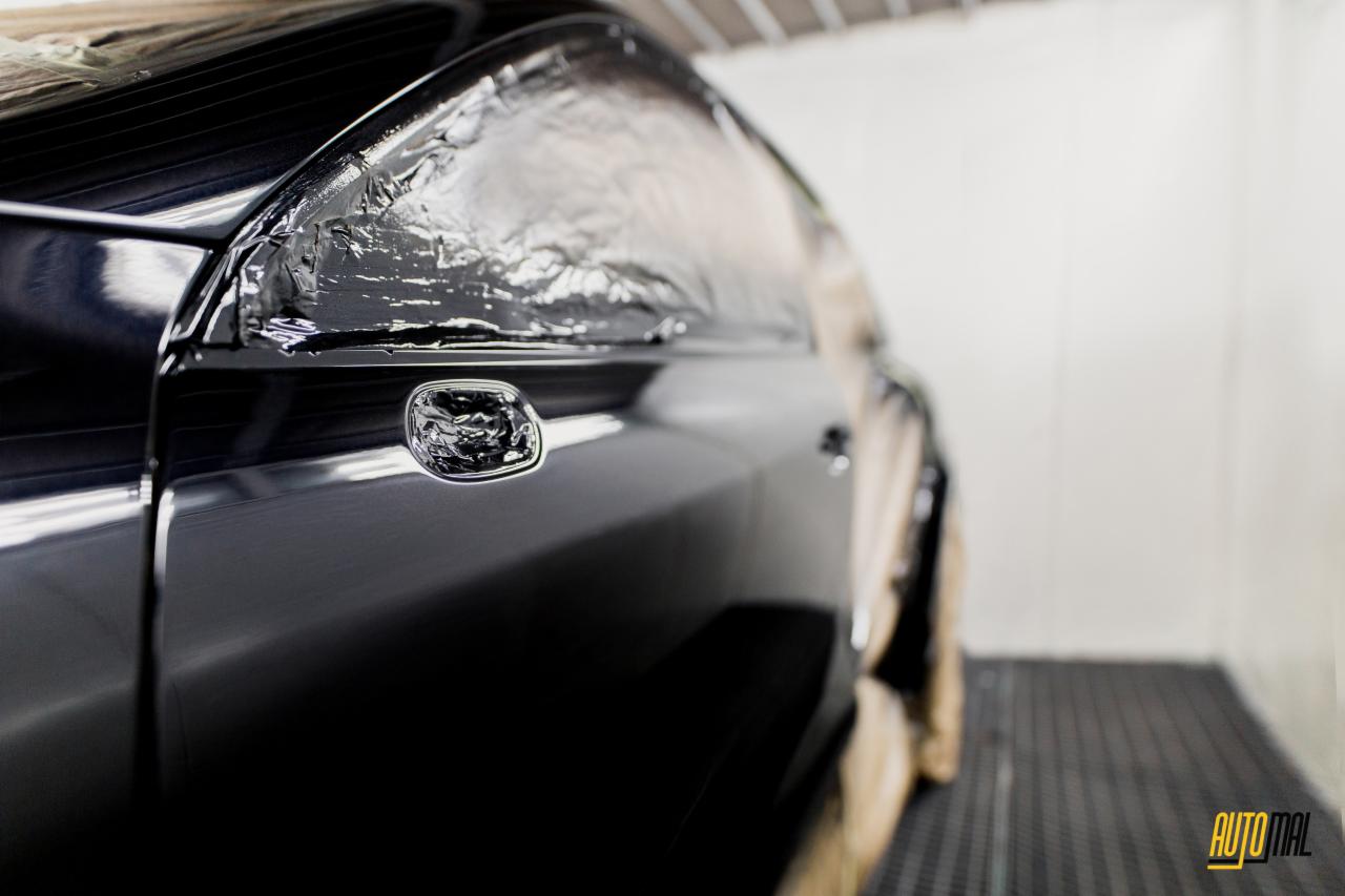 Błyszczący, czarny samochód częściowo oklejony folią ochronną w kabinie lakierniczej, widoczny chromowany uchwyt drzwi.