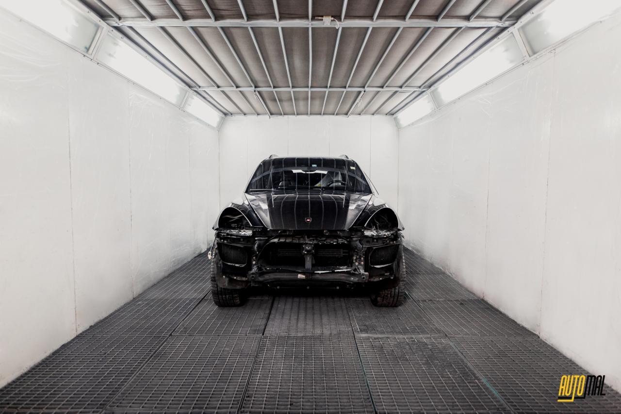 Czarny samochód osobowy, prawdopodobnie SUV, bez przedniego zderzaka i reflektorów, w jasnym pomieszczeniu z kratowaną podłogą, prawdopodobnie przygotowywany do naprawy karoserii.