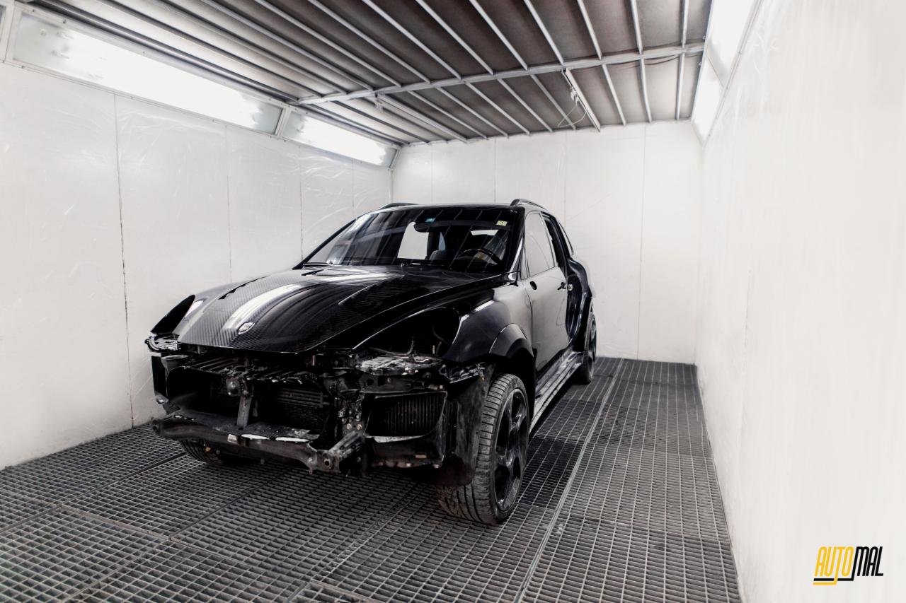 Czarny SUV w komorze lakierniczej, z widocznymi uszkodzeniami karoserii przed naprawą, na podłodze kratownica odprowadzająca farbę.