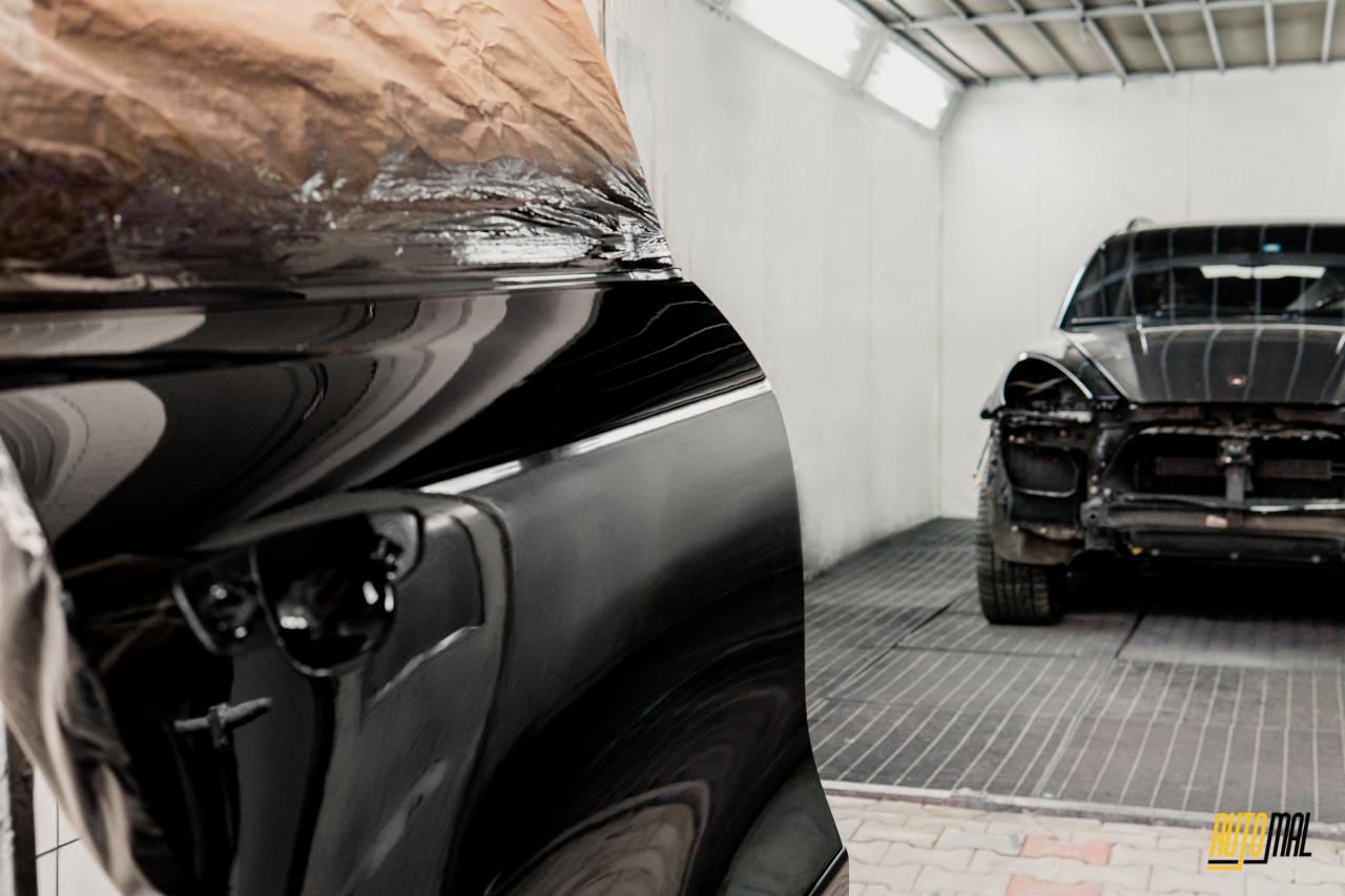 Fragment karoserii czarnego samochodu zabezpieczony papierem maskującym przed lakierowaniem, w tle drugi samochód po demontażu elementów karoserii w komorze lakierniczej.