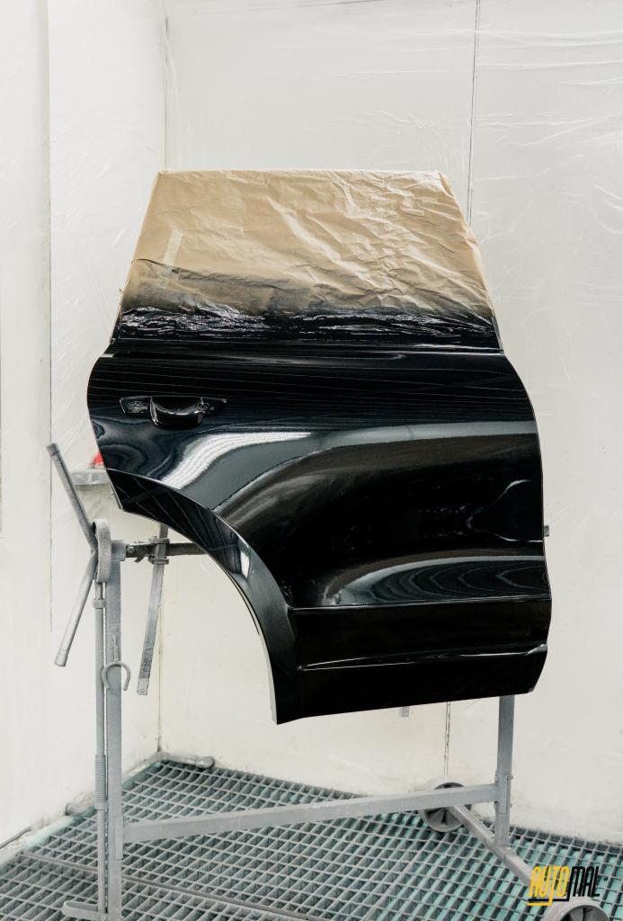 Czarna, błyszcząca karoseria drzwi samochodowych, częściowo zakryta papierem maskującym, zawieszona na stojaku w lakierni. Widoczna kratka podłogowa i logo firmy w prawym dolnym rogu.