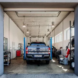 Szary Ford Ranger Wildtrak na podnośniku w jasnym warsztacie samochodowym, widok z tyłu, widoczne opony, narzędzia i wyposażenie warsztatu.