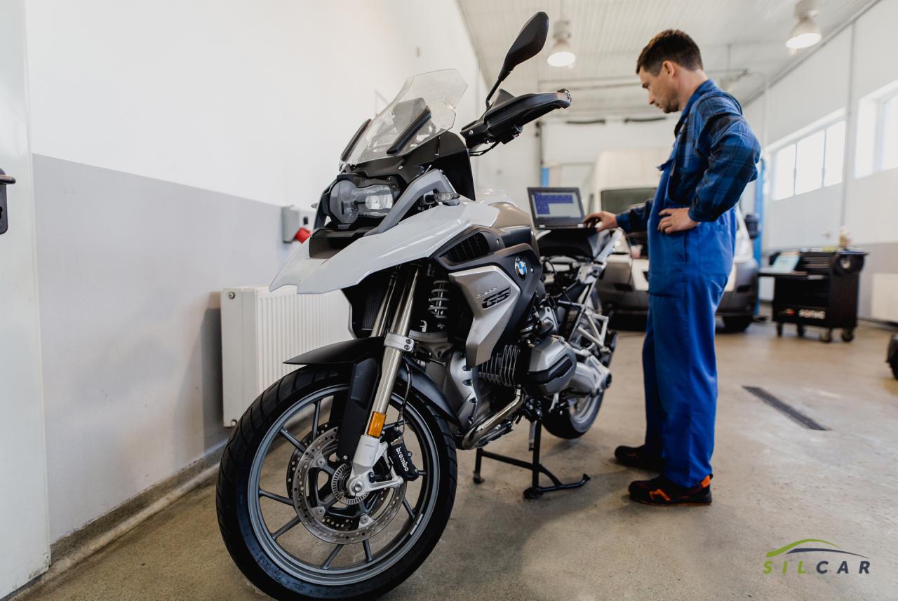 Biało-szary motocykl BMW GS na stojaku w warsztacie, mechanik w niebieskim kombinezonie diagnozuje go za pomocą laptopa. Widoczne narzędzia i wyposażenie warsztatu w tle.