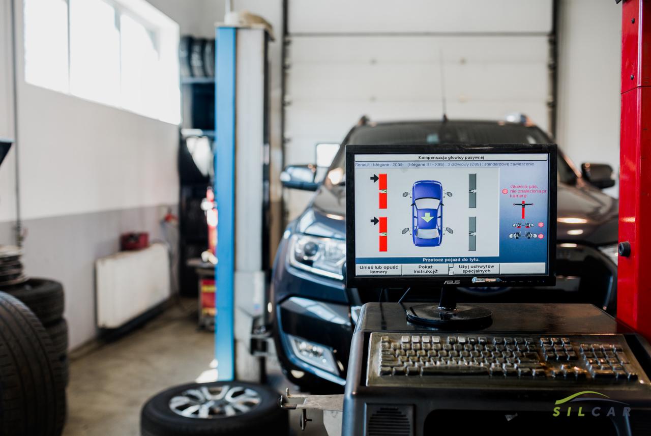 Samochód na stanowisku diagnostycznym z widocznym ekranem komputera prezentującego parametry ustawienia kół i osi; w tle opona i podnośnik samochodowy.