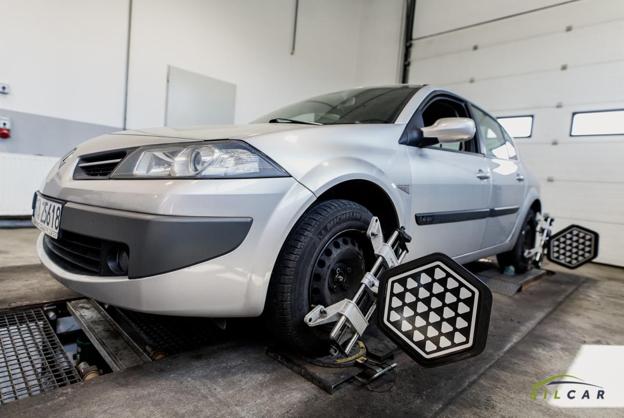 Srebrny samochód na stanowisku do ustawiania geometrii kół w warsztacie samochodowym, z widocznymi tarczami pomiarowymi na kołach.