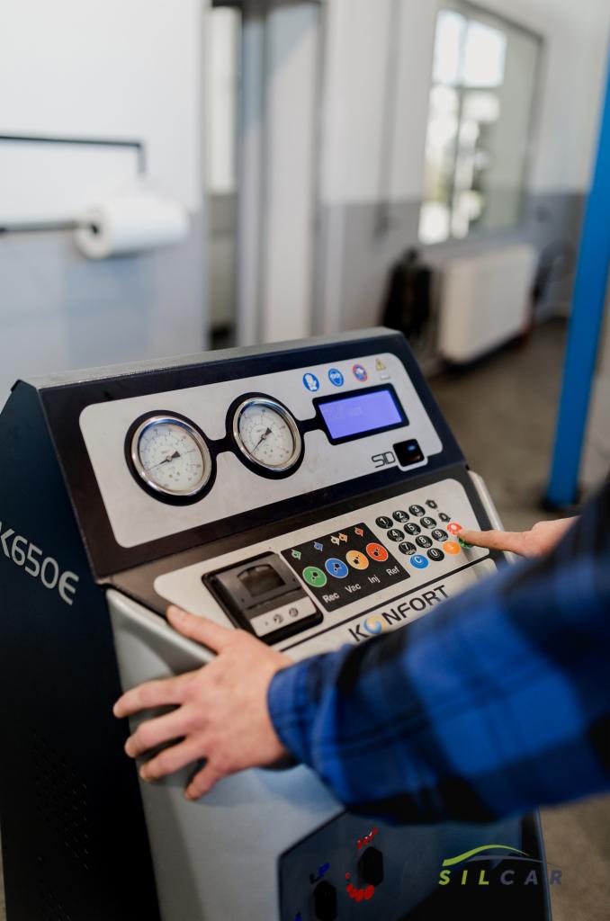 Obsługa klimatyzacji samochodowej: palec naciska przycisk na panelu sterowania urządzenia do serwisowania klimatyzacji, widoczne manometry i wyświetlacz, w tle warsztat samochodowy.