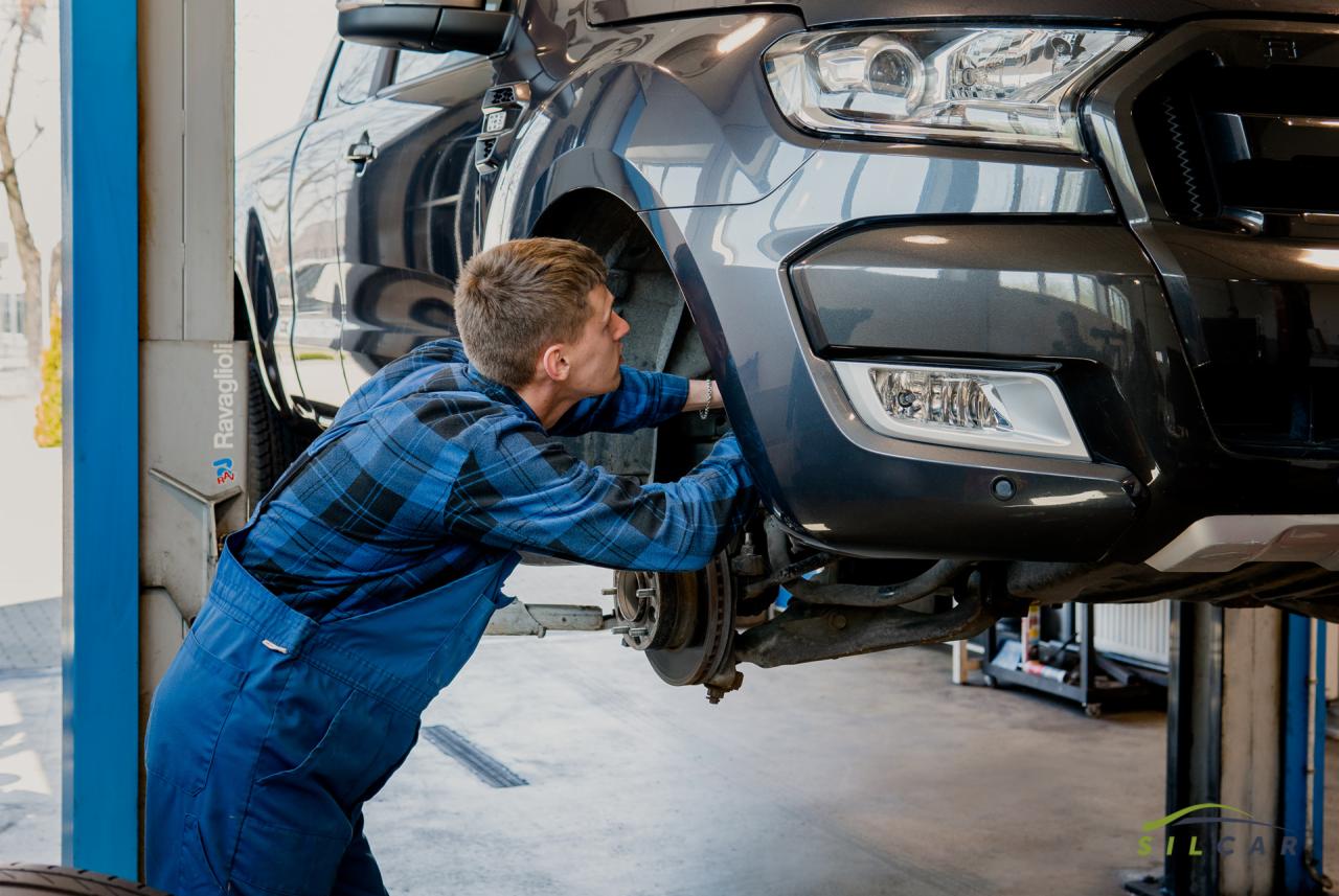 Mechanik w niebieskim kombinezonie i kraciastej koszuli pracuje przy zawieszeniu szarego pickupa na podnośniku, przednie koło zdjęte.