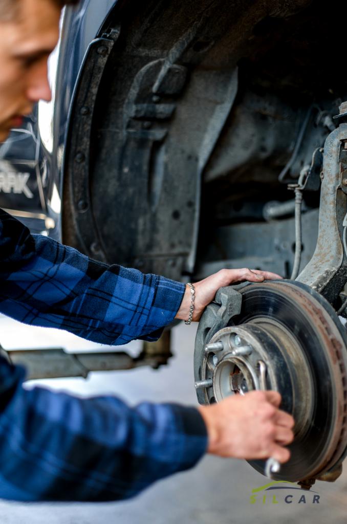 Mechanik w niebieskiej koszuli w kratę sprawdza tarczę hamulcową samochodu podniesionego na podnośniku, z widocznym kluczem w dłoni i srebrną bransoletką na nadgarstku.