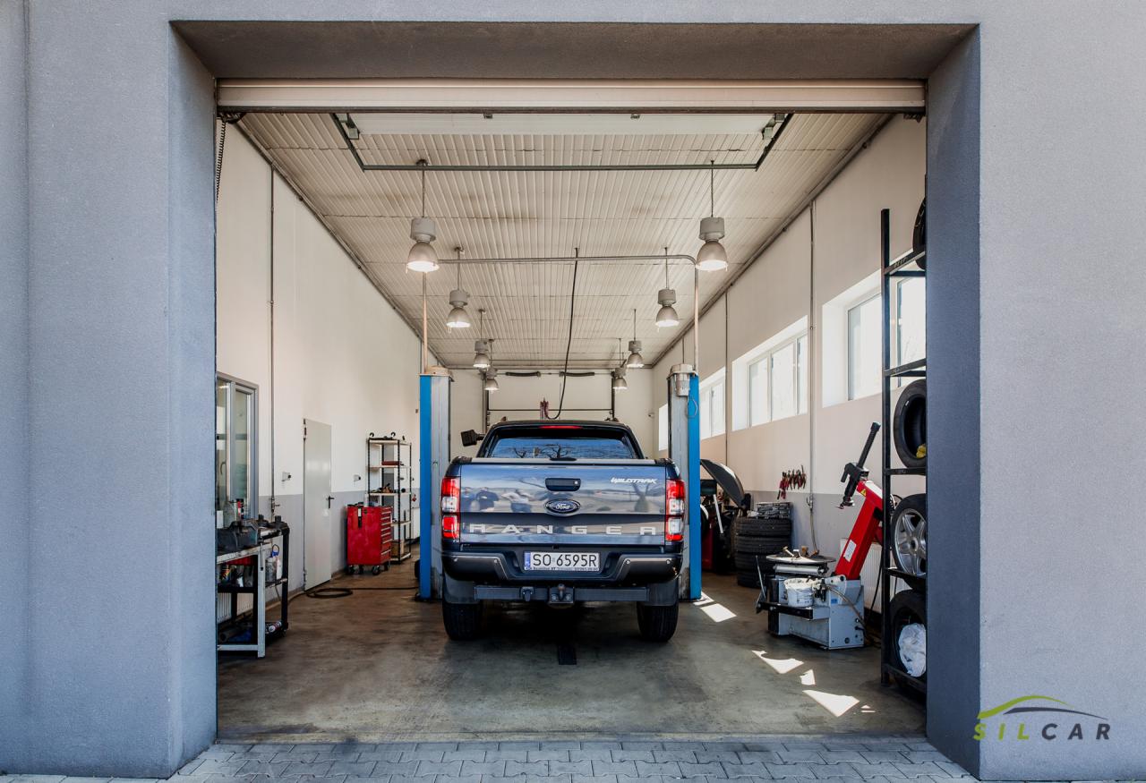 Szary Ford Ranger Wildtrak na podnośniku w jasnym warsztacie samochodowym, widok z tyłu, widoczne opony, narzędzia i wyposażenie warsztatu.