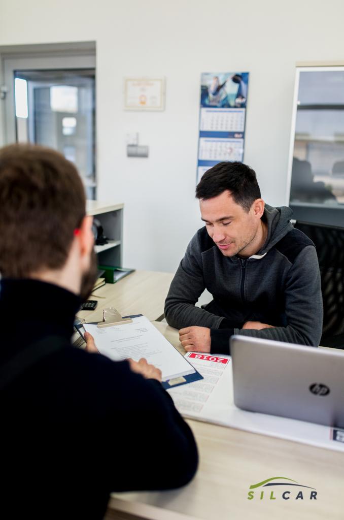 Mężczyzna w szarej bluzie przegląda dokumenty na biurku w warsztacie samochodowym, klient po lewej stronie, laptop i kalendarz na biurku, logo warsztatu w rogu
