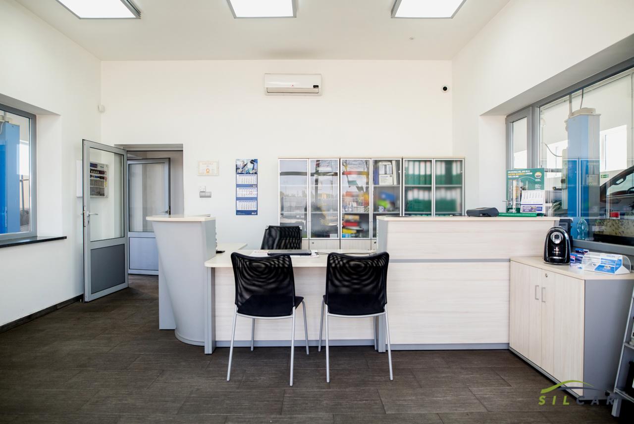 Jasne wnętrze recepcji warsztatu samochodowego z biurkiem, dwoma krzesłami, regałami z dokumentami i widokiem na stanowisko naprawcze za szybą.