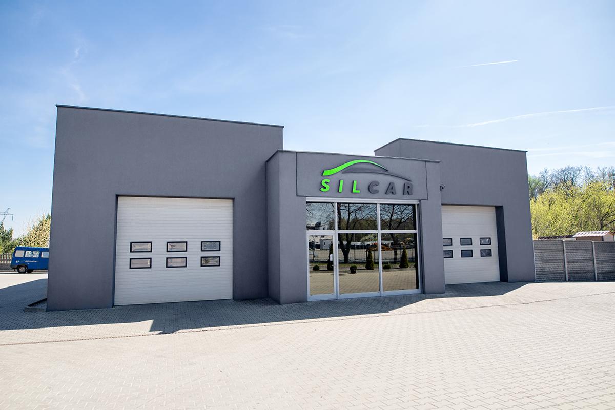 Nowoczesny budynek warsztatu samochodowego z szarymi ścianami, białymi bramami segmentowymi z przeszkleniami i dużymi szklanymi drzwiami wejściowymi z widocznym logo 'SILCAR' w kolorze zielonym.
