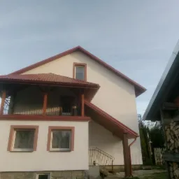 Dwupiętrowy dom z jasną elewacją i czerwonym dachem, balkon z balustradą, widoczne drewno na balkonie i schody z metalową poręczą, obok skład drewna.