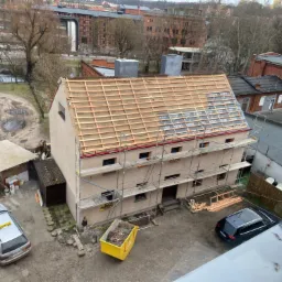 Dwupiętrowy budynek w trakcie prac dekarskich, widoczne rusztowania, przygotowana konstrukcja dachu z membraną, kontener na odpady, samochody na podwórzu, ujęcie z góry w pochmurny dzień.