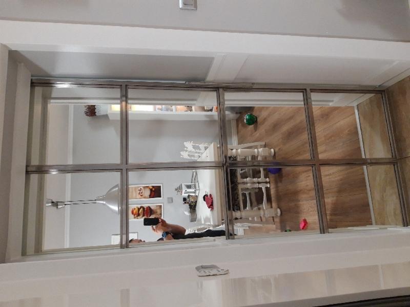 Wnętrze domu widziane przez przeszkloną balustradę z metalową ramą, odbicie osoby robiącej zdjęcie telefonem.