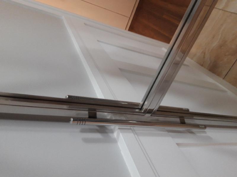 Szklana balustrada z nierdzewnymi profilami mocowana do białej ściany z drewnianym elementem dekoracyjnym na górze.