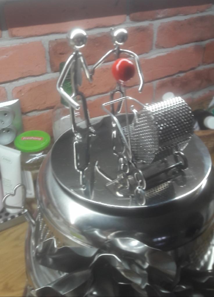 Metalowa figurka przedstawiająca dwie osoby pchające wózek z czerwonym elementem na tle ściany z czerwonej cegły, prawdopodobnie dekoracja wnętrza w Warszawie.