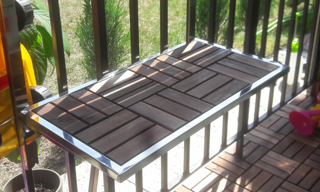 Balkon z drewnianą podłogą z płytek i metalową balustradą z drewnianym blatem, widok z góry, w tle zieleń.