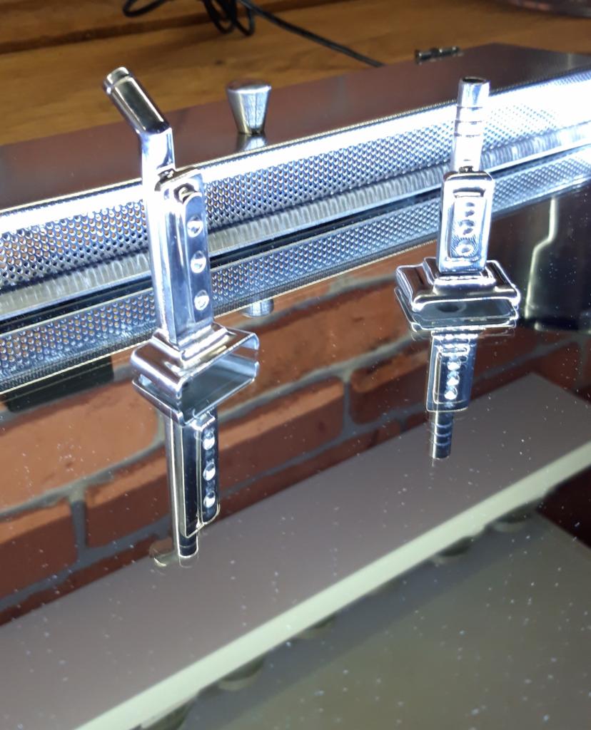 Dwa chromowane słupki mocujące, prawdopodobnie do balustrady lub poręczy, ustawione na błyszczącej powierzchni z odbiciem ceglanej ściany w tle.