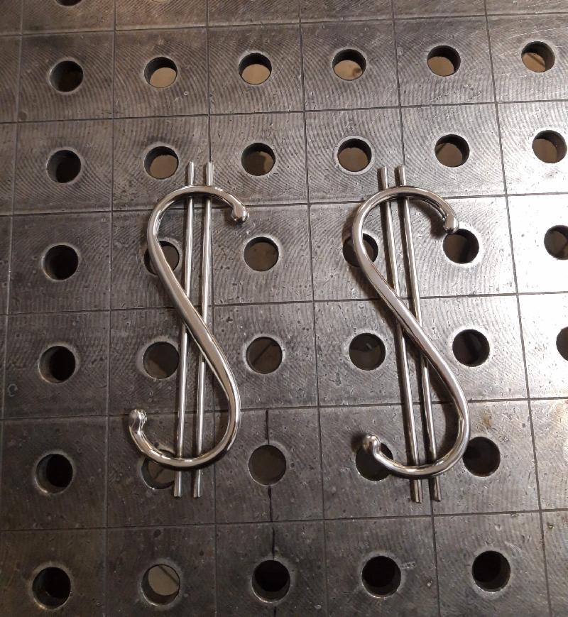 Dwa metalowe symbole dolara wykonane z polerowanych rurek, leżące na perforowanym metalowym stole spawalniczym.