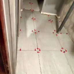 Szare płytki podłogowe w trakcie układania, z czerwonymi i niebieskimi klipsami poziomującymi, widoczne przy drzwiach i rurze grzewczej.