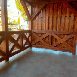 Zadaszony balkon z drewnianą balustradą w kratkę i podłogą wyłożoną terakotą, widok na zieleń i budynki w tle.