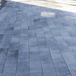 Ułożona szara kostka brukowa na podjeździe, z widoczną furtką z kutego żelaza w tle, kilka kostek jeszcze nie ułożonych.