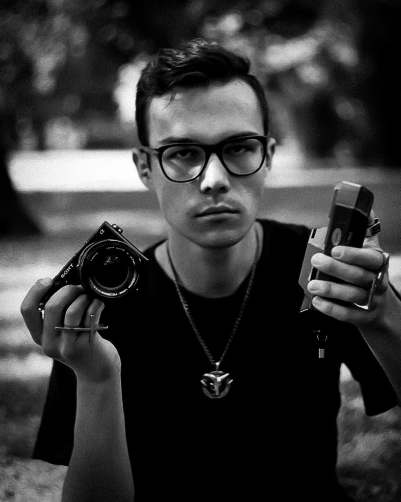 Czarno-białe zdjęcie fotografa z aparatem Sony i światłomierzem w dłoniach, na tle rozmytego parku. Mężczyzna w okularach i z naszyjnikiem patrzy w obiektyw.