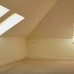 Poddasze z oknami dachowymi i małym oknem ściennym, ściany w jasnym odcieniu beżu, widoczne gniazdka elektryczne, surowe wykończenie podłogi, drzwi po prawej stronie.