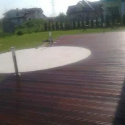 Nowoczesny taras z ciemnego drewna z okrągłym, betonowym jacuzzi wbudowanym w podest, w tle dom jednorodzinny i trawnik.