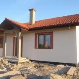 Nowo wybudowany, parterowy dom jednorodzinny z czerwonym dachem i jasną elewacją, widoczny komin i drewniane wykończenia ganku, otoczony ziemią i resztkami materiałów budowlanych.