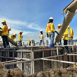 Pol Krusz - Wylewanie betonu do szalunku fundamentów przez grupę pracowników w odblaskowych kamizelkach, widok z dołu na tle błękitnego nieba.