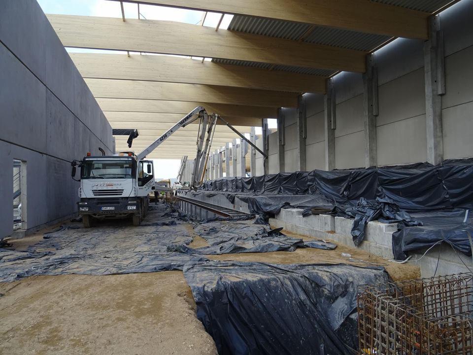 Betonowanie fundamentów hali produkcyjnej z użyciem pompy do betonu; widoczne szalunki zabezpieczone folią, zbrojenie i drewniana konstrukcja dachu.