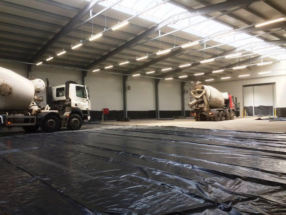 Dwa samochody ciężarowe z betonomieszarkami w hali w Kruklinie, podłoga wyłożona czarną folią, widoczne oświetlenie sufitowe.