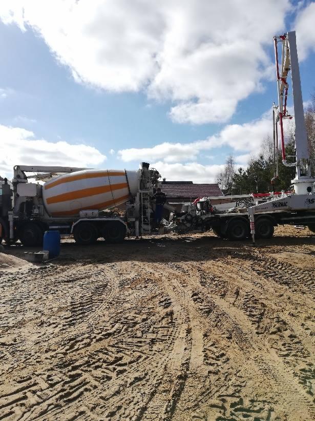 Ciężarówka z betonomieszarką i pompą do betonu na placu budowy, widoczne ślady opon na piasku, błękitne niebo z chmurami w tle.