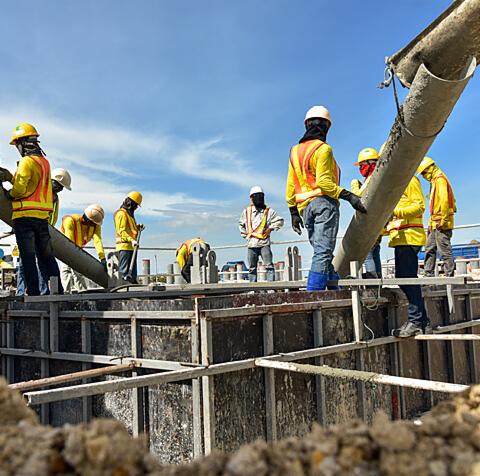 Wylewanie betonu do szalunku fundamentów przez grupę pracowników w odblaskowych kamizelkach, widok z dołu na tle błękitnego nieba.