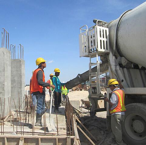Pracownicy w kaskach i kamizelkach odblaskowych nadzorują wylewanie betonu z betonomieszarki na placu budowy z widocznymi zbrojeniami i szalunkami.
