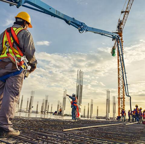Budowa w toku: robotnicy w kaskach i kamizelkach odblaskowych na zbrojeniu, widoczny dźwig z pompą do betonu i pionowe słupy, słoneczne oświetlenie.