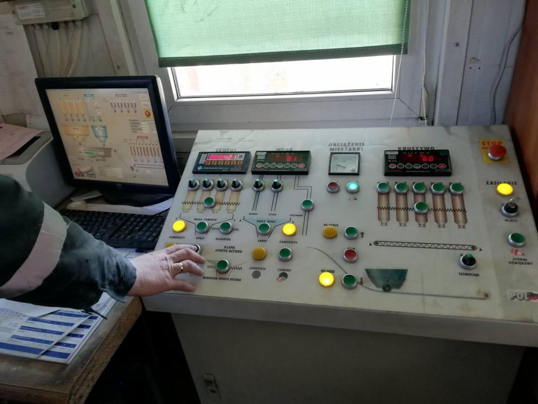 Panel sterowania betoniarnią z włączonymi wskaźnikami cementu, wody i kruszywa; widoczny monitor z wizualizacją procesu mieszania betonu oraz dłoń operatora wciskająca przycisk.