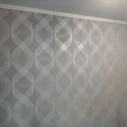 Ściana pokryta szarą tapetą z geometrycznym wzorem w odcieniach bieli, widoczny fragment drabiny malarskiej w prawym dolnym rogu.